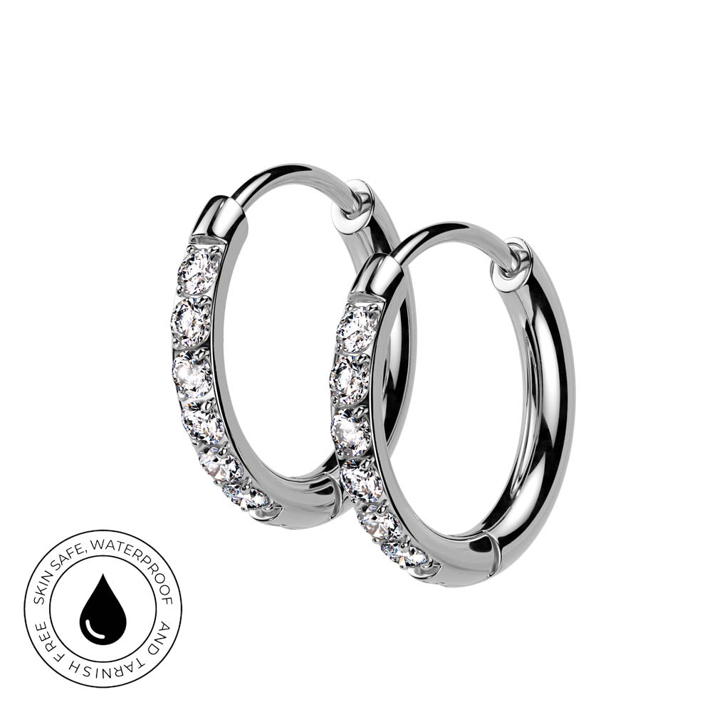 A pair of silver hoop earrings with clear pavé-set stones on one side, shown on a white background with a circular 'Skin Safe Waterproof' badge in the lower-left corner.