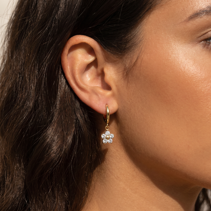 A close-up of a model's ear wearing a small gold hoop earring with a dangling clear crystal flower charm.