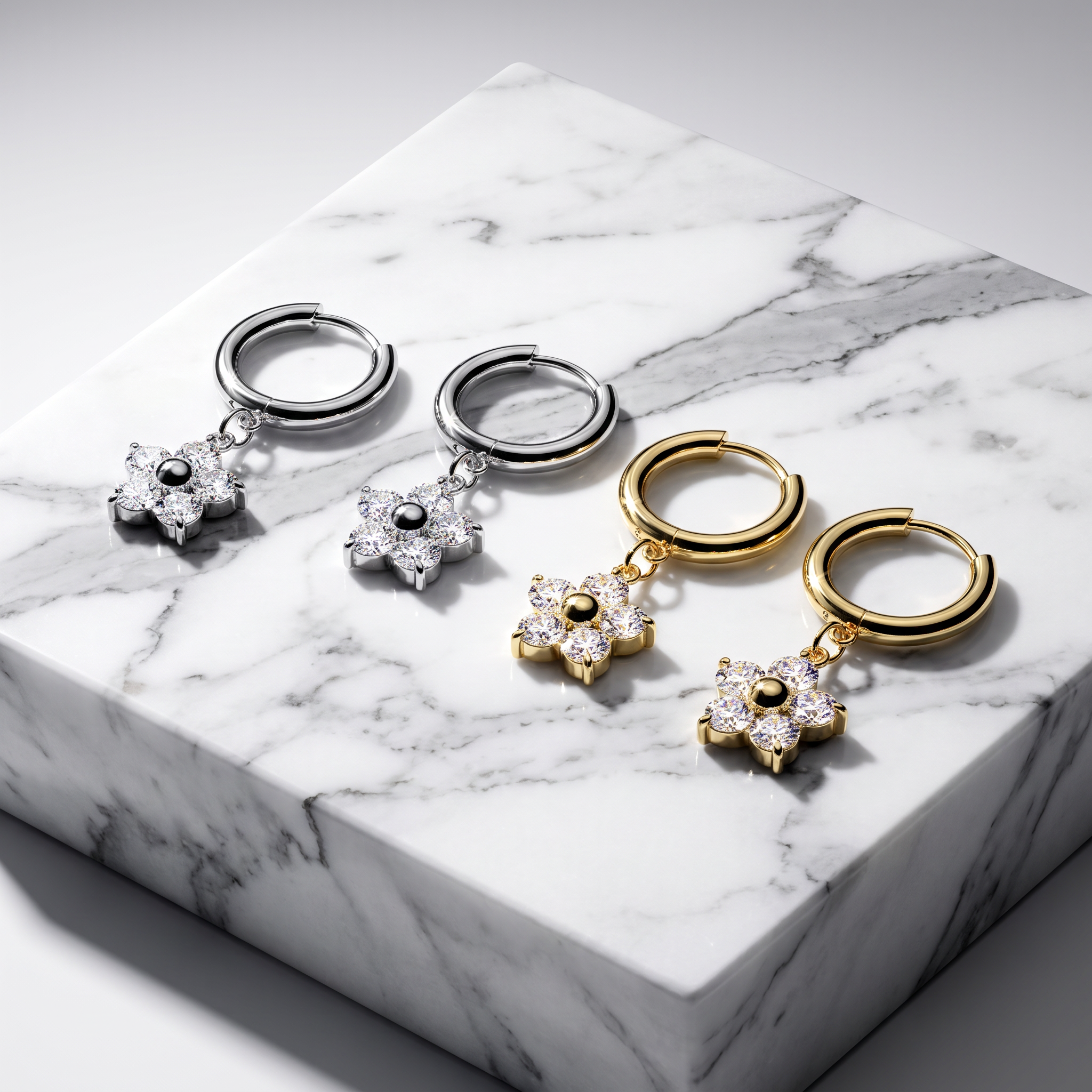 Four small hoop earrings with dangling flower charms are arranged on a white marble block. Two pairs are silver-toned and two are gold-toned, each with clear stones in the flower center.