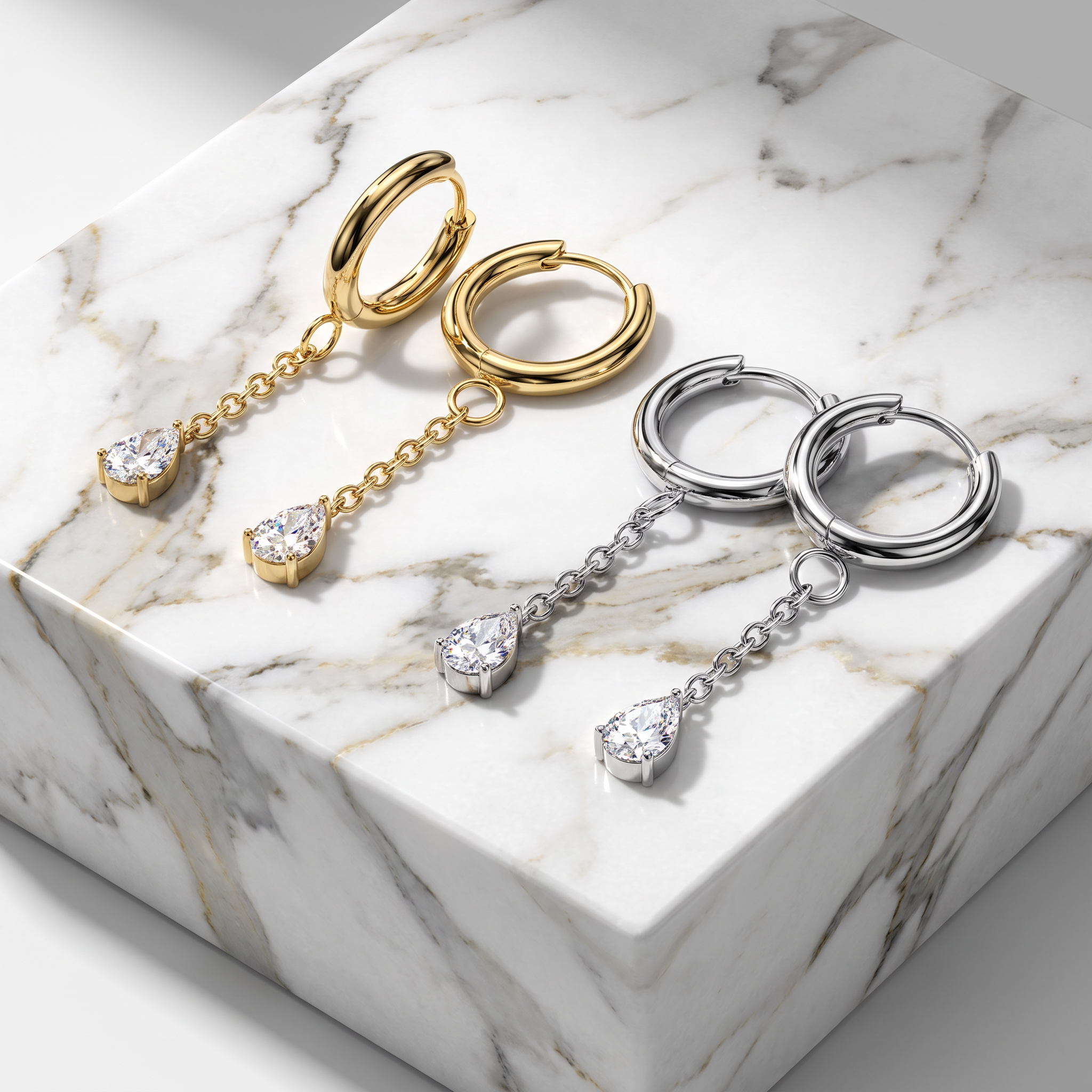 Two pairs of dangling hoop earrings displayed on a white marble block. One pair is yellow gold with a small teardrop-shaped clear gemstone at the bottom of each hoop and chain.