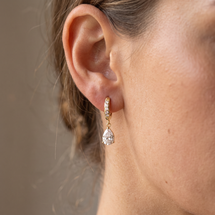 A close-up of a person's ear wearing a small gold huggie hoop earring adorned with clear stones and a dangling pear-cut clear gemstone.