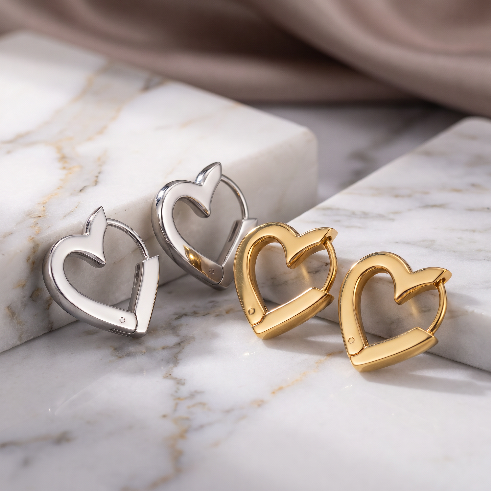 Four heart-shaped hoop earrings in silver and gold finishes, arranged on a white marble surface with a soft beige fabric in the background.