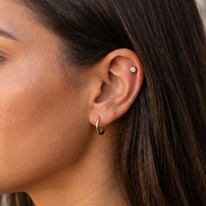 Close-up of a person's ear wearing a small gold hoop earring and a tiny round stud earring, with dark brown hair and warm skin tones.