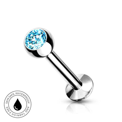 A silver-toned labret piercing barbell with a round bezel-set light blue faceted gemstone, shown on a white background with a small circular 'Skin Safe Waterproof' badge in the lower-left corner.