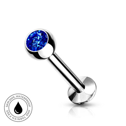 A silver-toned labret piercing barbell with a round, faceted blue gemstone on the end, shown on a clean white background with a small circular 'Skin Safe Waterproof' badge in the lower-left corner.
