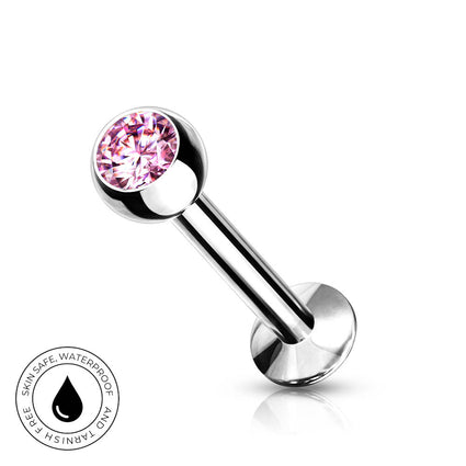 A silver-toned labret piercing barbell with a faceted pink gemstone ball end, shown on a clean white background with a small circular skin-safe waterproof badge in the lower left corner.
