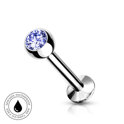 A silver-toned labret piercing barbell with a round blue faceted gemstone on the end, shown on a clean white background with a small circular skin-safe waterproof badge in the lower left corner.