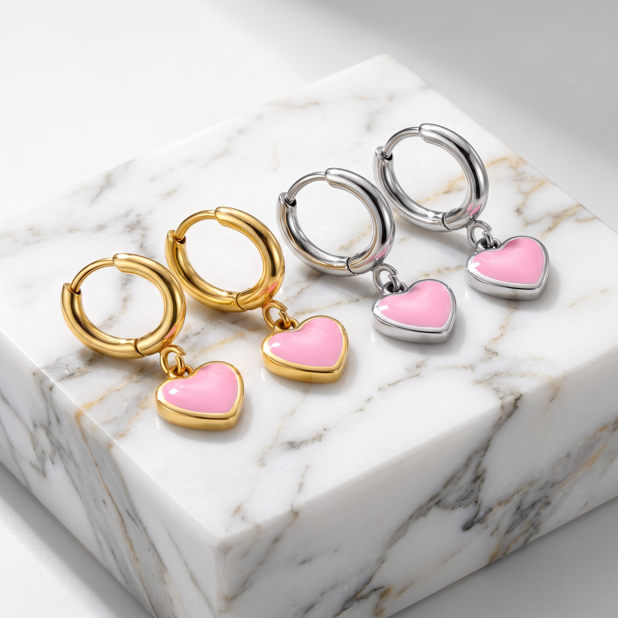 Three pairs of hoop earrings with dangling heart charms are arranged on a white marble block. The left pair is gold-toned with a pink heart charm, the middle pair is silver-toned with a pink heart charm, and the right pair has matching silver-toned hoops with a pink heart charm.