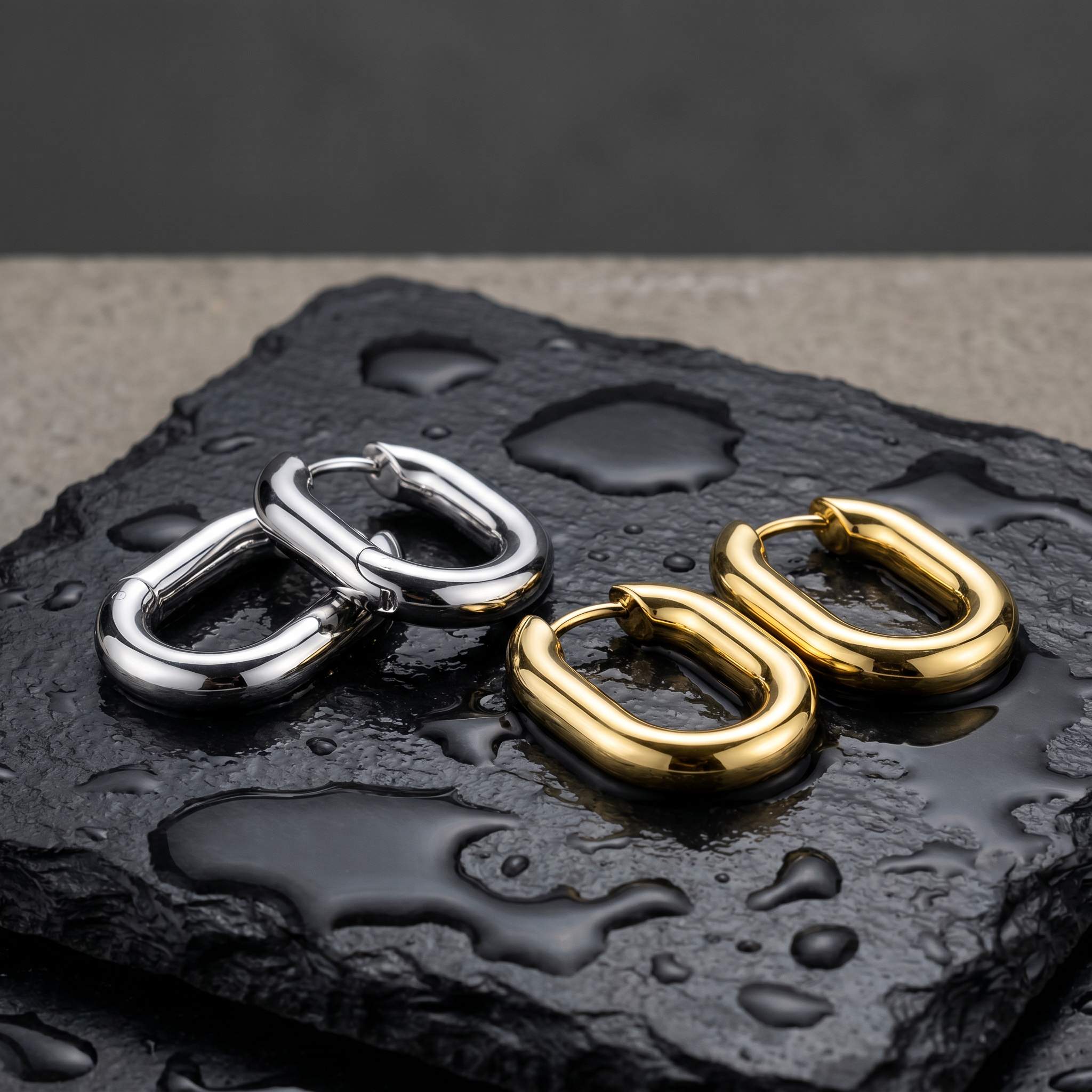 Two pairs of chunky geometric hoop earrings, one in polished silver and one in polished gold, displayed on a wet dark slate stone surface.