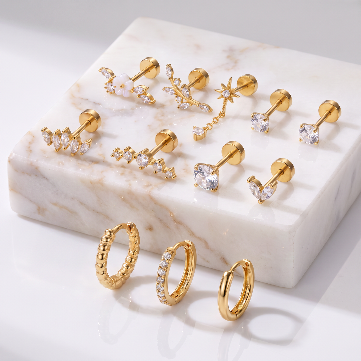 A set of gold-toned stud earrings and hoop earrings displayed on a white marble block against a clean white background. The earrings include clear crystal accents in various shapes such as moons, stars, and curved lines, with two textured hoop earrings and two huggie hoop earrings shown in front.