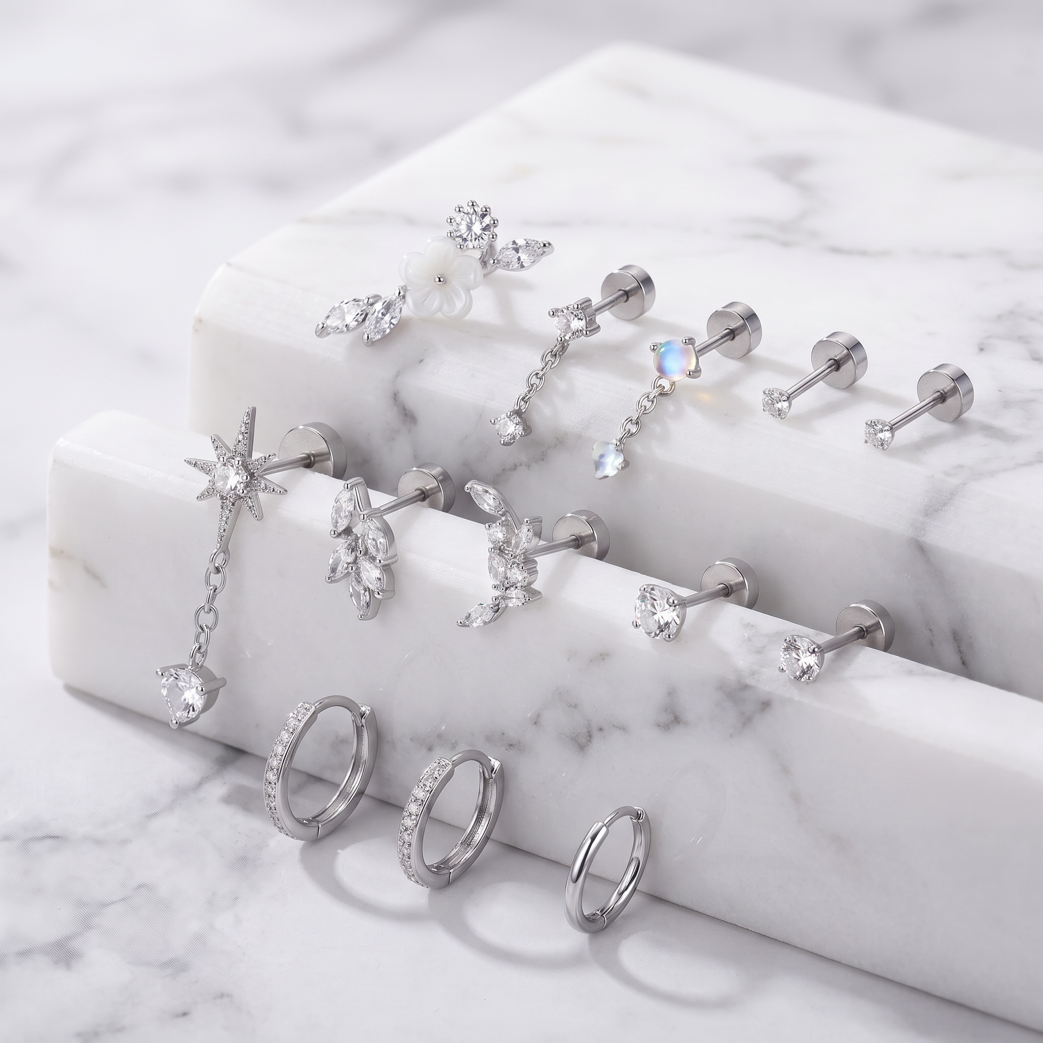 A styled product shot of multiple silver-toned ear piercings and rings displayed on white marble blocks over a marble surface. The jewelry includes hoop earrings, studs, and dangling charms with clear stones in floral, starburst, and marquise shapes.