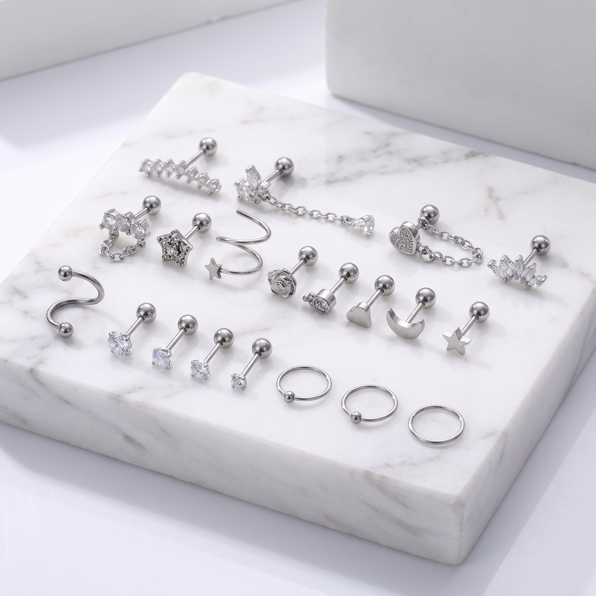 A set of silver-toned body jewelry pieces arranged on a white marble display block, including studs, rings, and curved barbell-style earrings with clear stones and metallic charms.