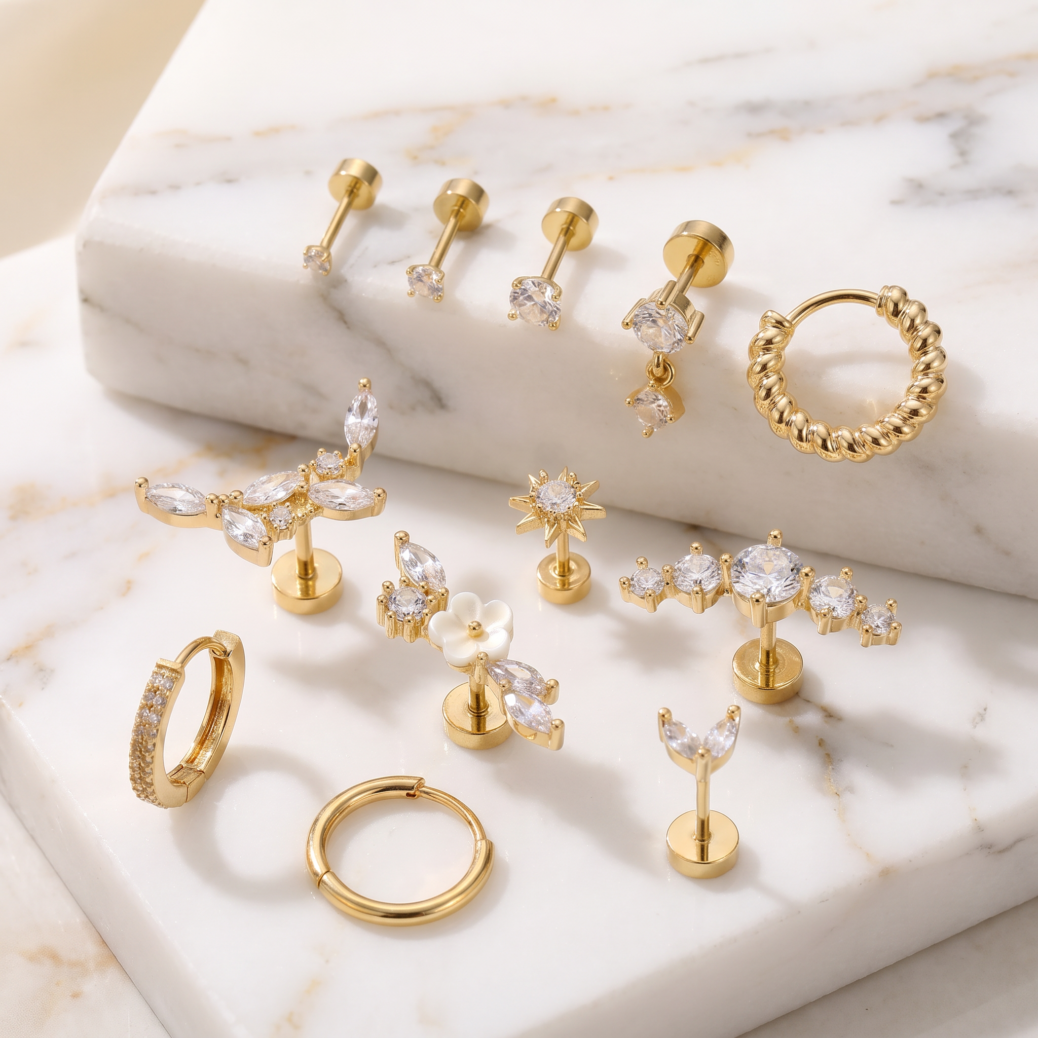 A set of gold-toned body jewelry and earrings arranged on white marble display blocks. The collection includes multiple hoop, stud, and cartilage piercings in various shapes, with several pieces featuring clear crystal or cubic zirconia stones.