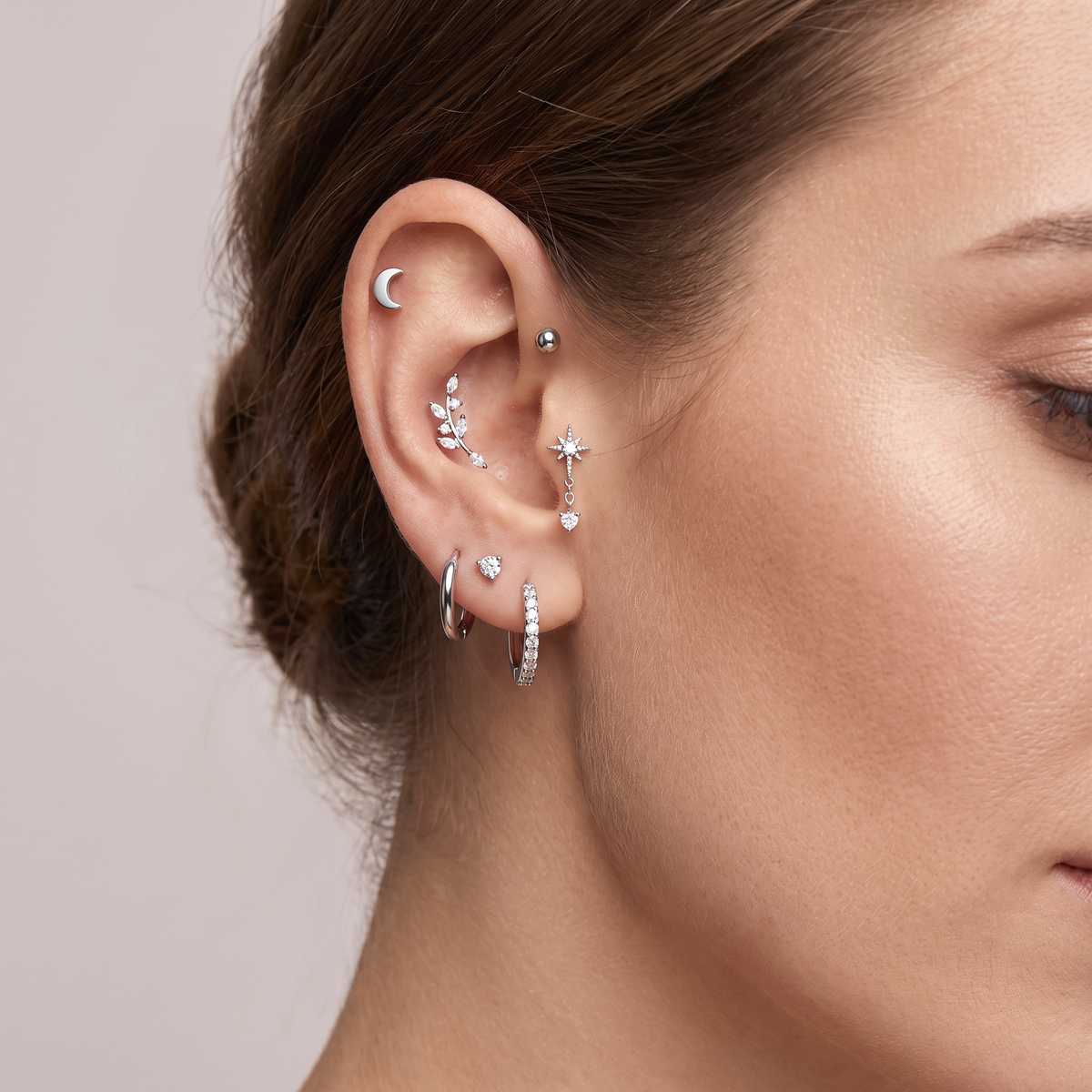 A close-up of a model's ear showcasing multiple silver-toned ear piercings, including a crescent moon stud, a cluster of small floral studs, a dangling star charm, a huggie hoop, and a pavé huggie hoop, against a soft beige background.