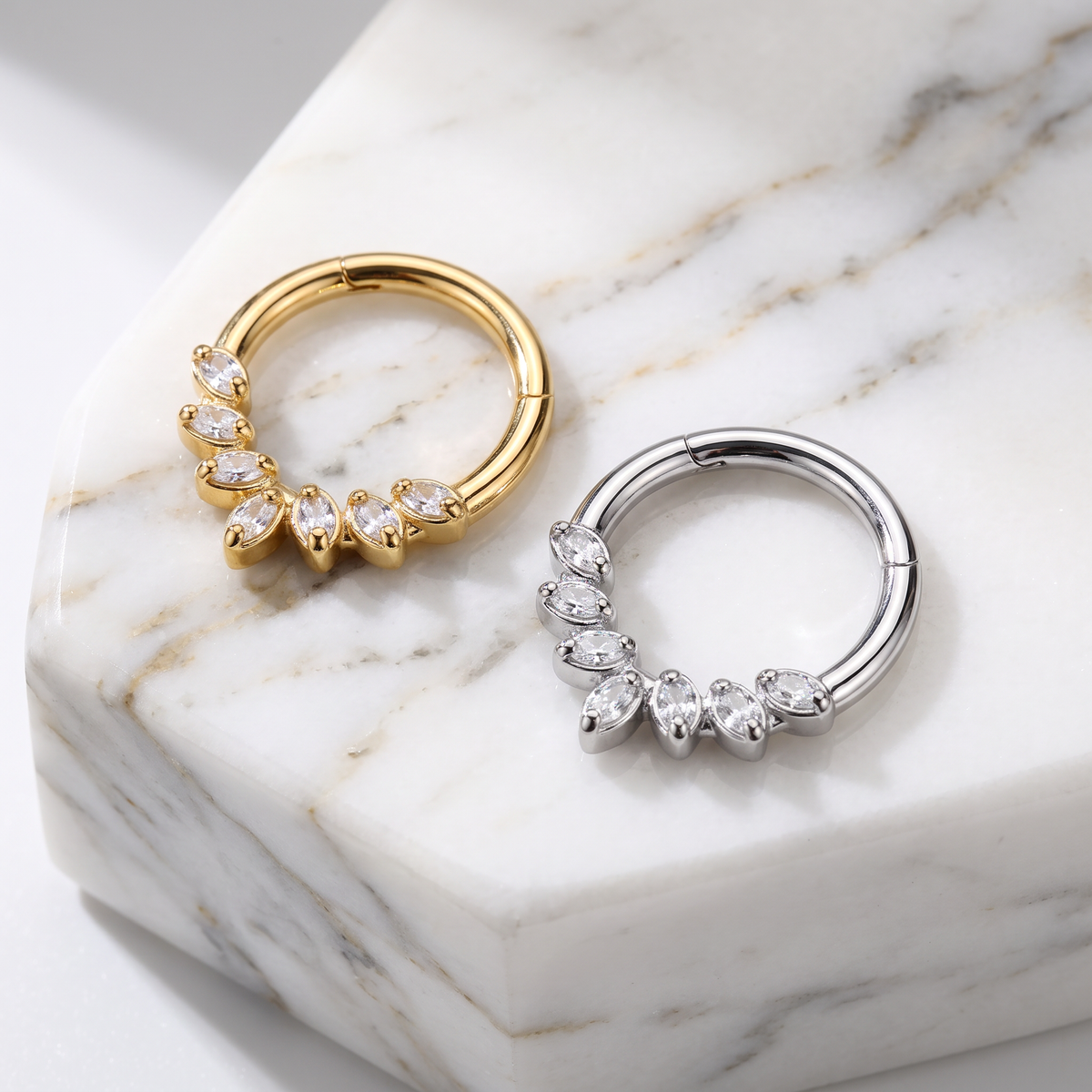 Two hoop earrings with marquise-cut clear crystals are displayed on a white marble block. One earring has a gold-tone metal body, and the other has a silver-tone metal body.