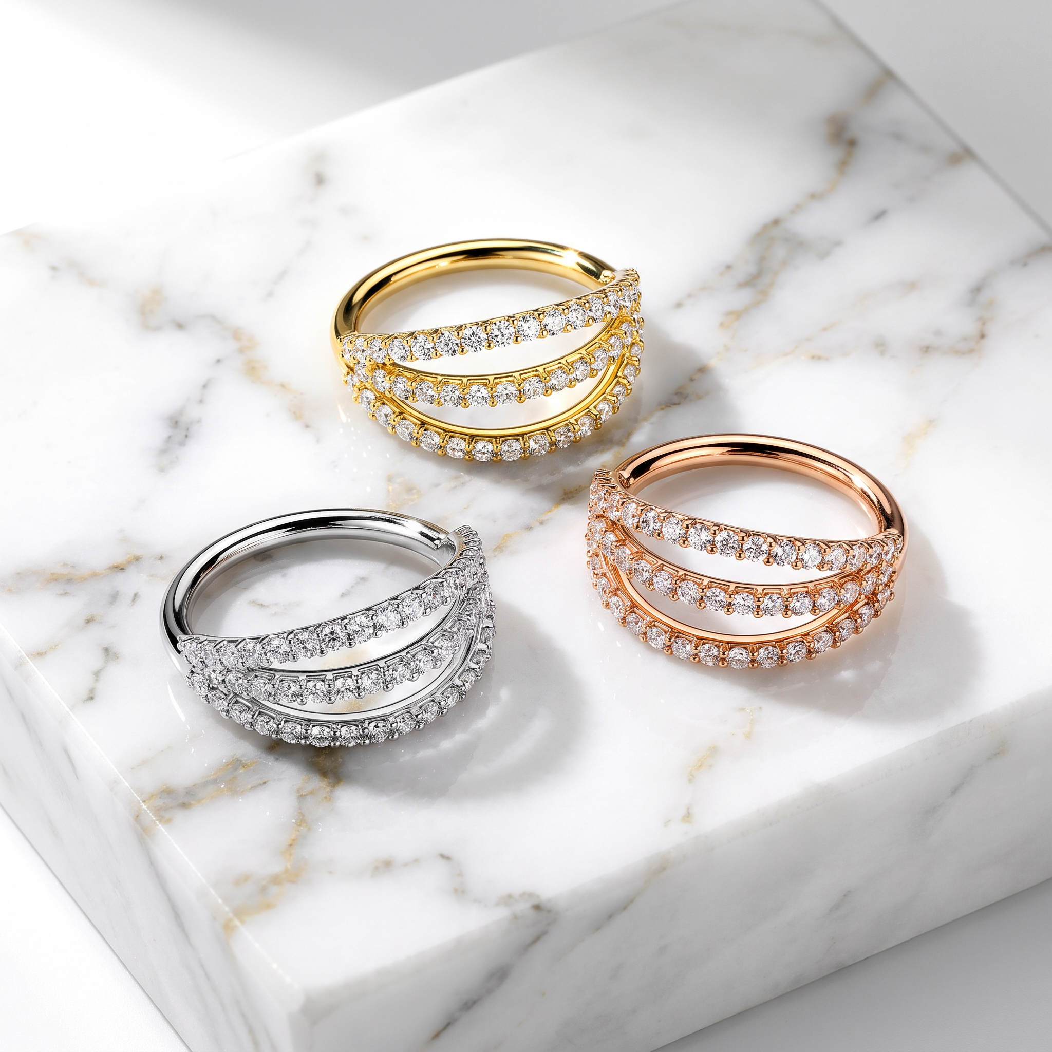 Three double-band wrap rings with pavé-set clear stones, shown in yellow gold, rose gold, and silver-tone metal on a white marble slab.