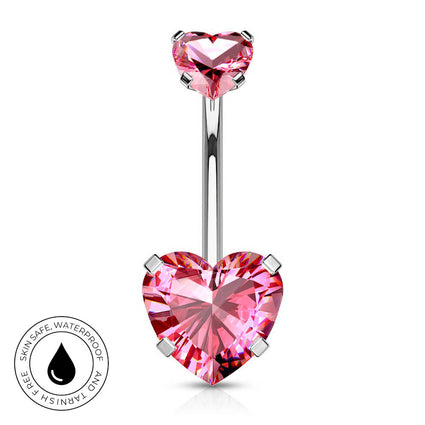 A silver-toned navel barbell piercing with a large, faceted pink heart-shaped crystal at the bottom and a smaller matching pink heart at the top. A circular badge in the bottom-left corner highlights skin-safe, waterproof quality.