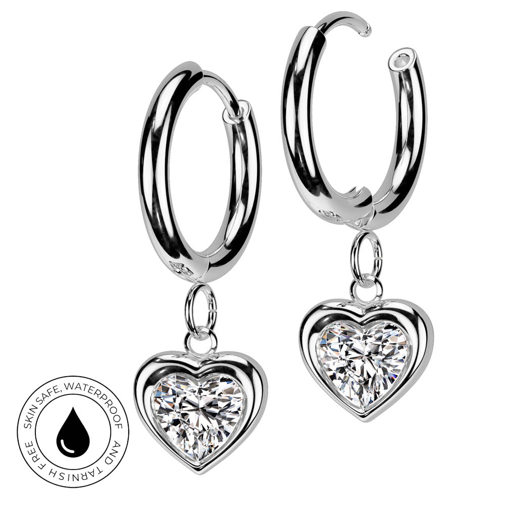 A pair of silver hoop earrings with dangling heart-shaped clear gemstone charms, shown on a white background with a circular skin-safe waterproof badge in the lower left corner.