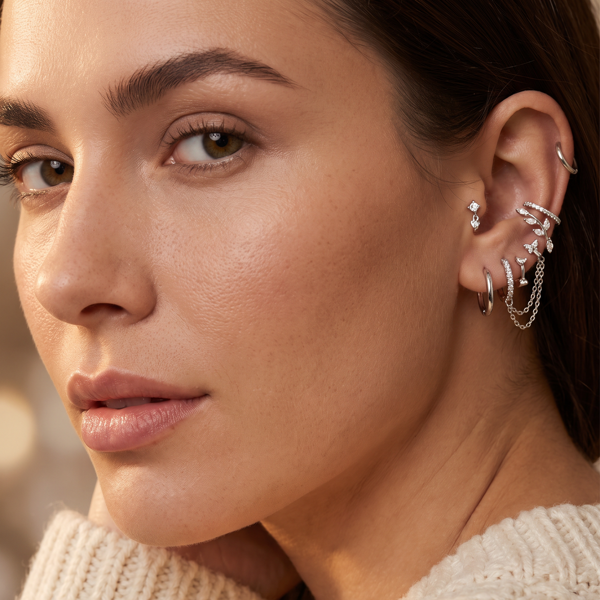 A close-up of a woman's ear adorned with multiple silver-toned ear cuffs and hoop earrings, including a curved ear cuff, a chain drop, and small hoop earrings. She is wearing a cream-colored knit sweater against a warm, softly blurred background.