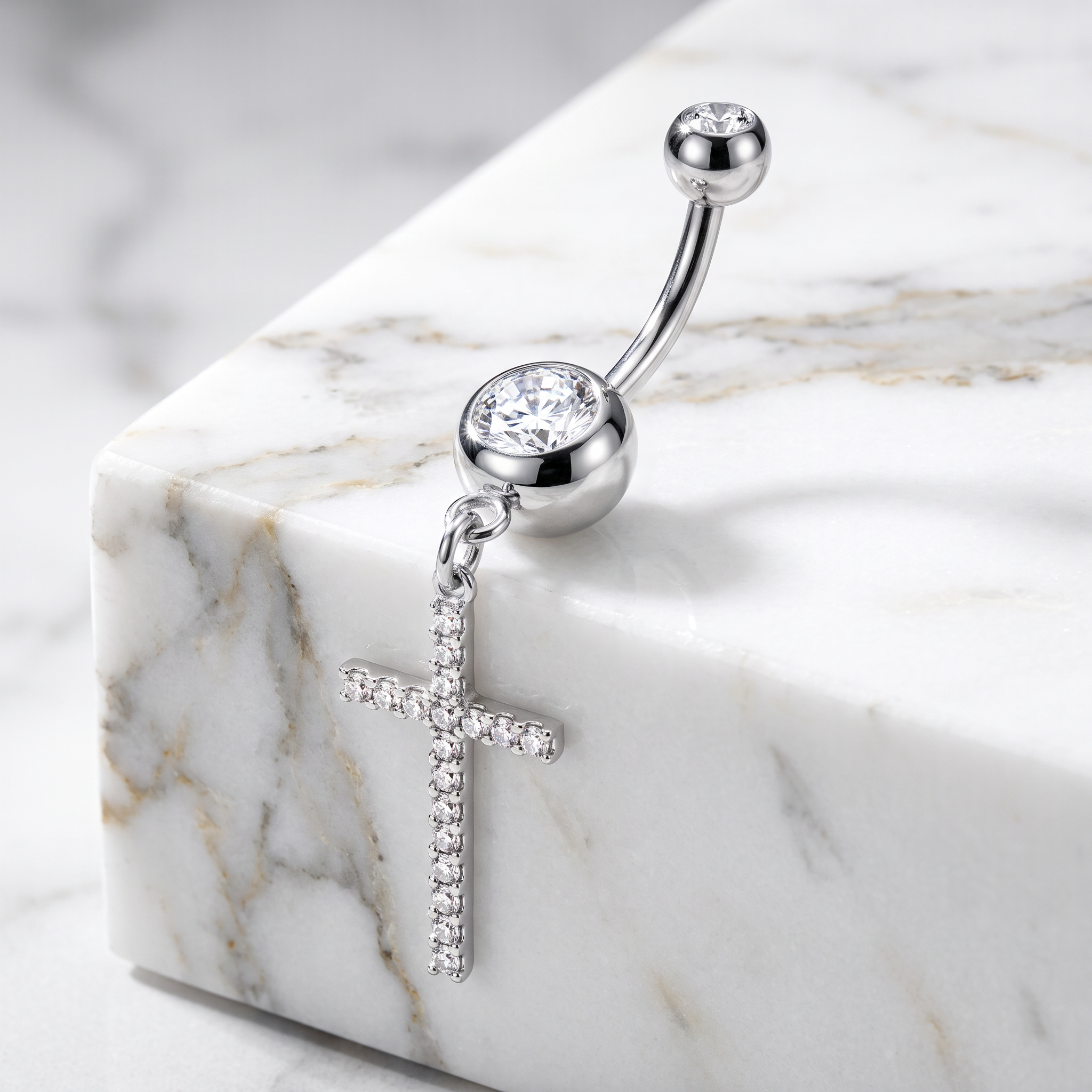 A silver-toned navel piercing ring with a curved barbell, a round bezel-set clear crystal, and a dangling cross charm adorned with small clear crystals, displayed on a white marble block.