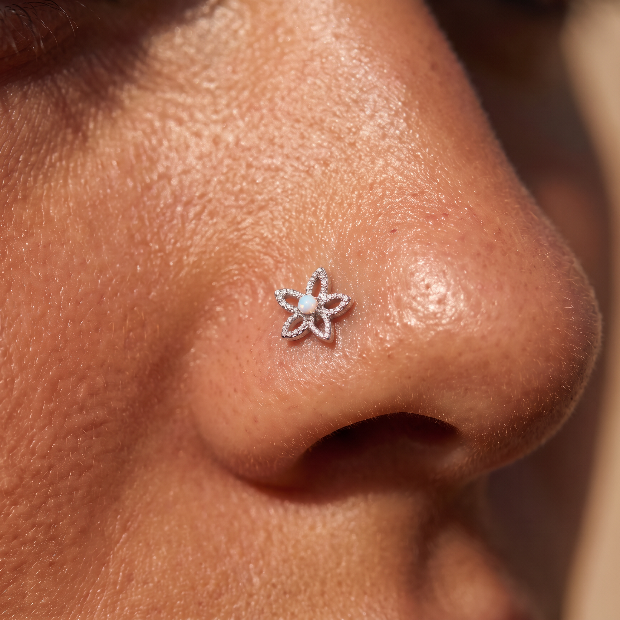 A close-up of a person's nose wearing a small silver flower-shaped nose stud with a clear central stone.