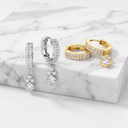 A set of gold and silver huggie hoop earrings embellished with pavé stones and dangling clear crystal charms, displayed on a white marble block.