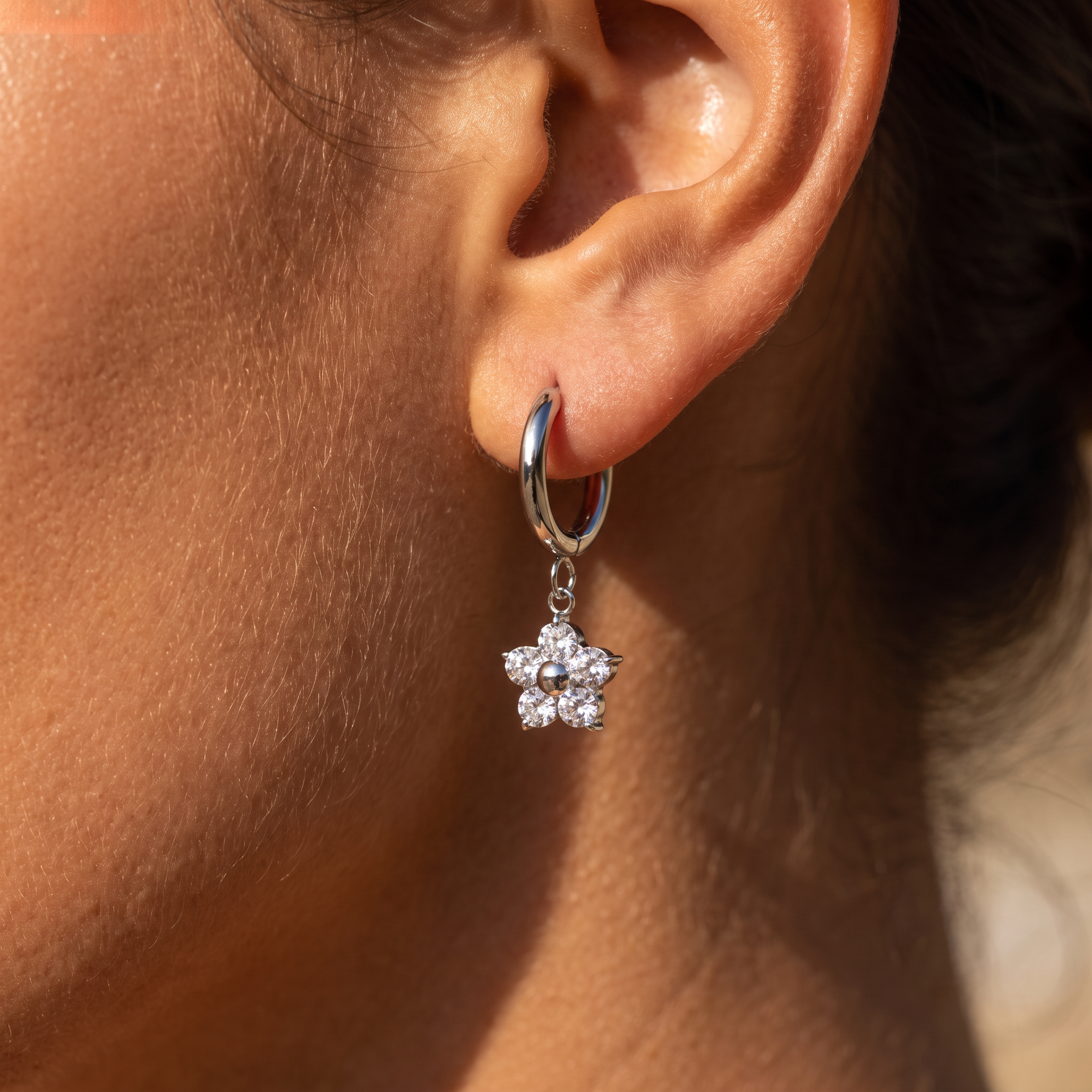 A close-up of a person's ear wearing a silver huggie hoop earring with a dangling flower charm made of clear stones.