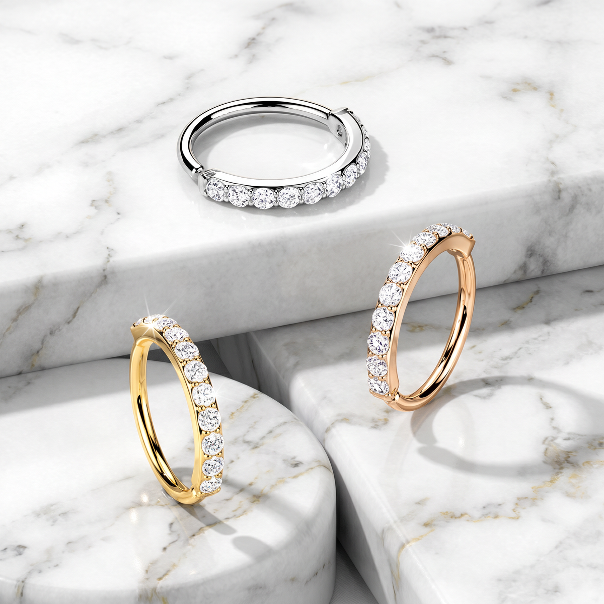 Three diamond hoop rings in silver, yellow gold, and rose gold, arranged on a white and gray marble pedestal background.