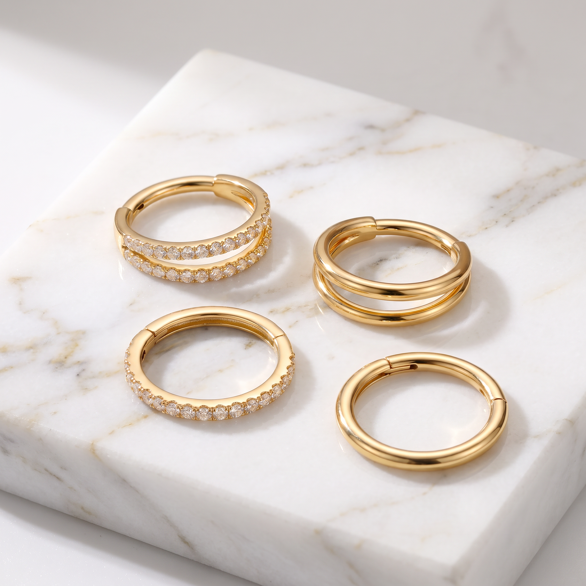 Four gold hoop earrings displayed on a white marble slab. Two hoops are adorned with small clear stones, and the remaining two are solid gold.