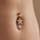 A close-up of a gold belly button ring with two dangling flower charms set with clear stones, worn on a person's navel.