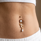 A close-up of a person's midsection wearing a gold belly button ring with clear rhinestone accents, styled with a white ribbed crop top and white pants.
