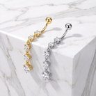 Two curved barbell belly button piercings with dangling diamond clusters, one in yellow gold and one in silver, displayed on a white marble block.