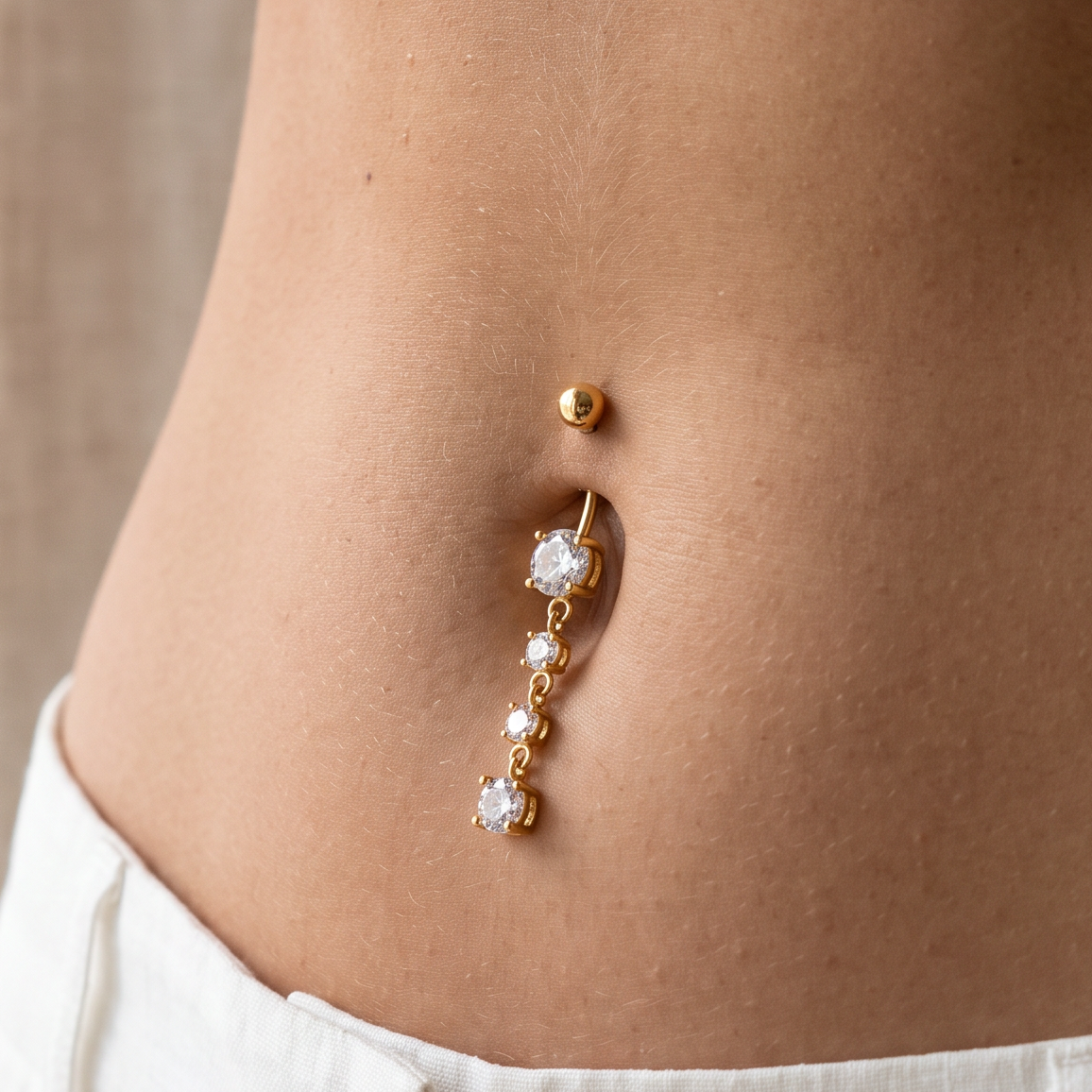 A close-up of a person's lower torso wearing a gold belly button ring with a dangling charm featuring clear rhinestones.