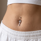 A close-up of a person's lower torso wearing a silver belly button ring with a heart-shaped dangling charm and a small silver dot stud above it, styled with a white ribbed crop top and white pants.