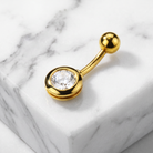 A gold-plated curved barbell navel piercing stud with a round bezel-set clear gemstone, displayed on a white marble block.