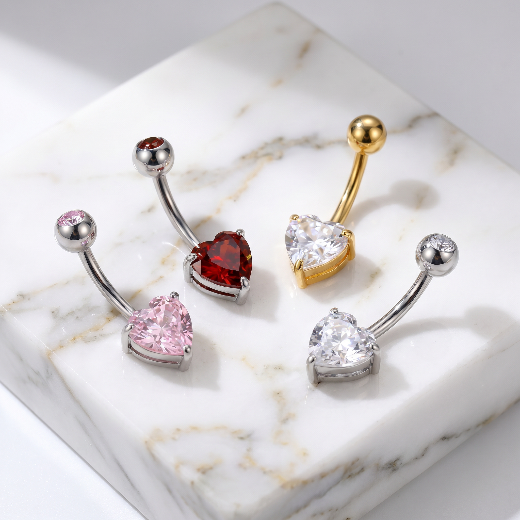 Four heart-shaped navel piercings displayed on a white marble block. The set includes silver-toned pieces with pink and clear stones, a red heart-shaped stone, and a gold-toned piece with a clear heart-shaped stone.