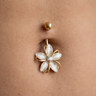 A close-up of a belly button piercing with a gold flower-shaped navel ring featuring white petal inlays and a small clear center stone, worn on skin.