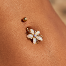 A gold-plated flower-shaped navel piercing with iridescent white opal-like petals and a small blue center stone, shown on a person's skin with a separate gold stud above it.