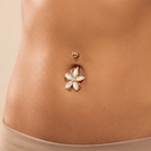 A close-up of a person's lower torso showing a gold-plated belly button ring with a flower-shaped charm featuring white petal inlays and a small light blue center, with a separate gold stud above it.