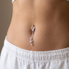 A close-up of a person's lower torso wearing a dangling belly button ring with clear rhinestones and a rose gold-toned metal setting, paired with white shorts and a white ribbed crop top.