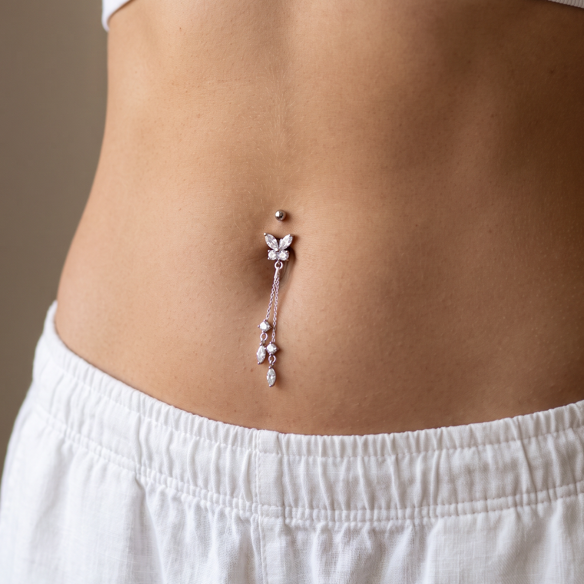 A close-up of a person's lower torso wearing a dangling belly button ring with clear rhinestones and a rose gold-toned metal setting, paired with white shorts and a white ribbed crop top.
