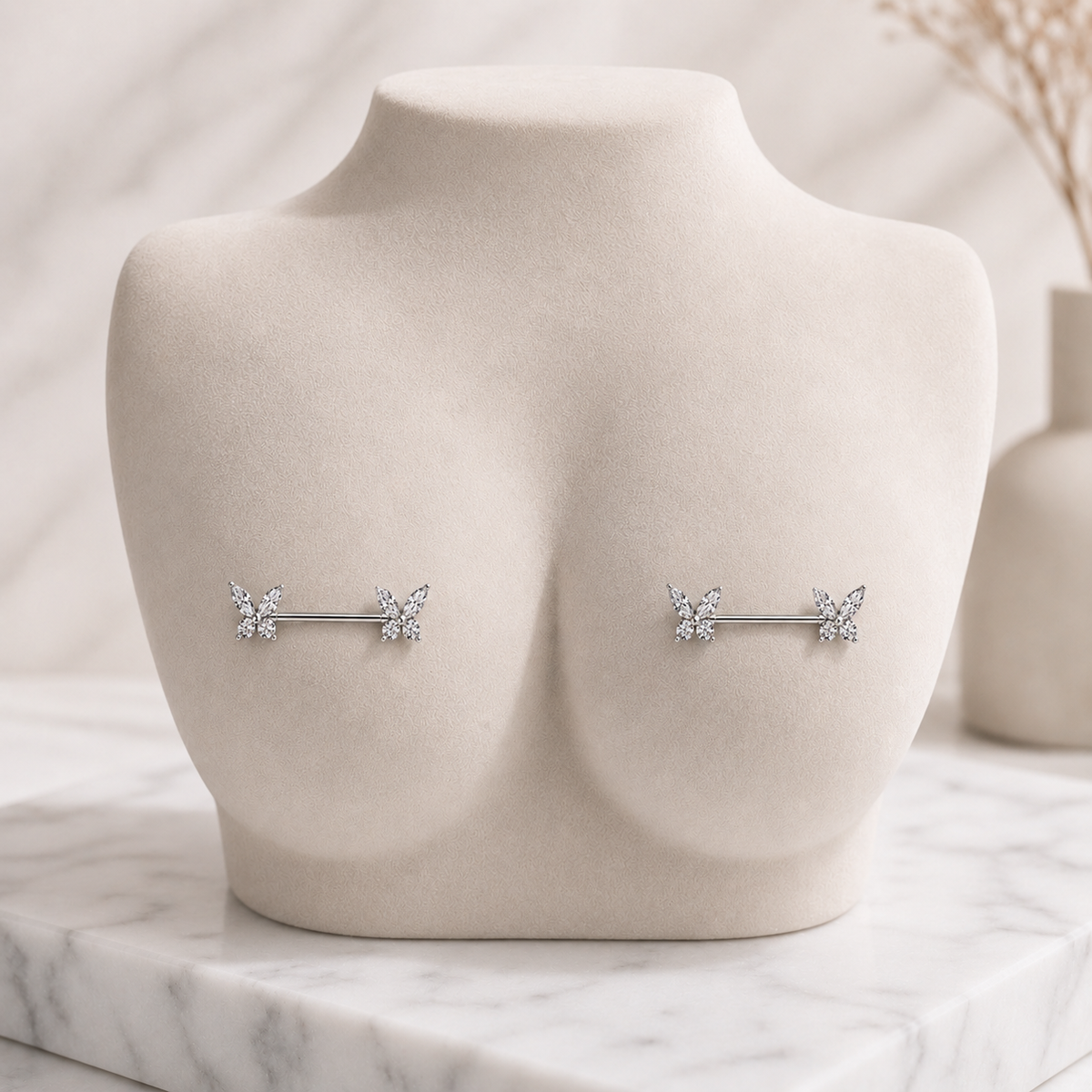 A pair of silver-toned barbell nipple piercings with small butterfly motifs, displayed on a beige jewelry bust against a soft marble and neutral background.