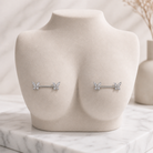 A pair of silver-toned barbell nipple piercings with small butterfly motifs, displayed on a beige jewelry bust against a soft marble and neutral background.