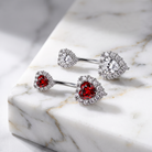 Three silver-toned belly button rings with heart-shaped red gemstones and clear halo stones, arranged on a white marble block.