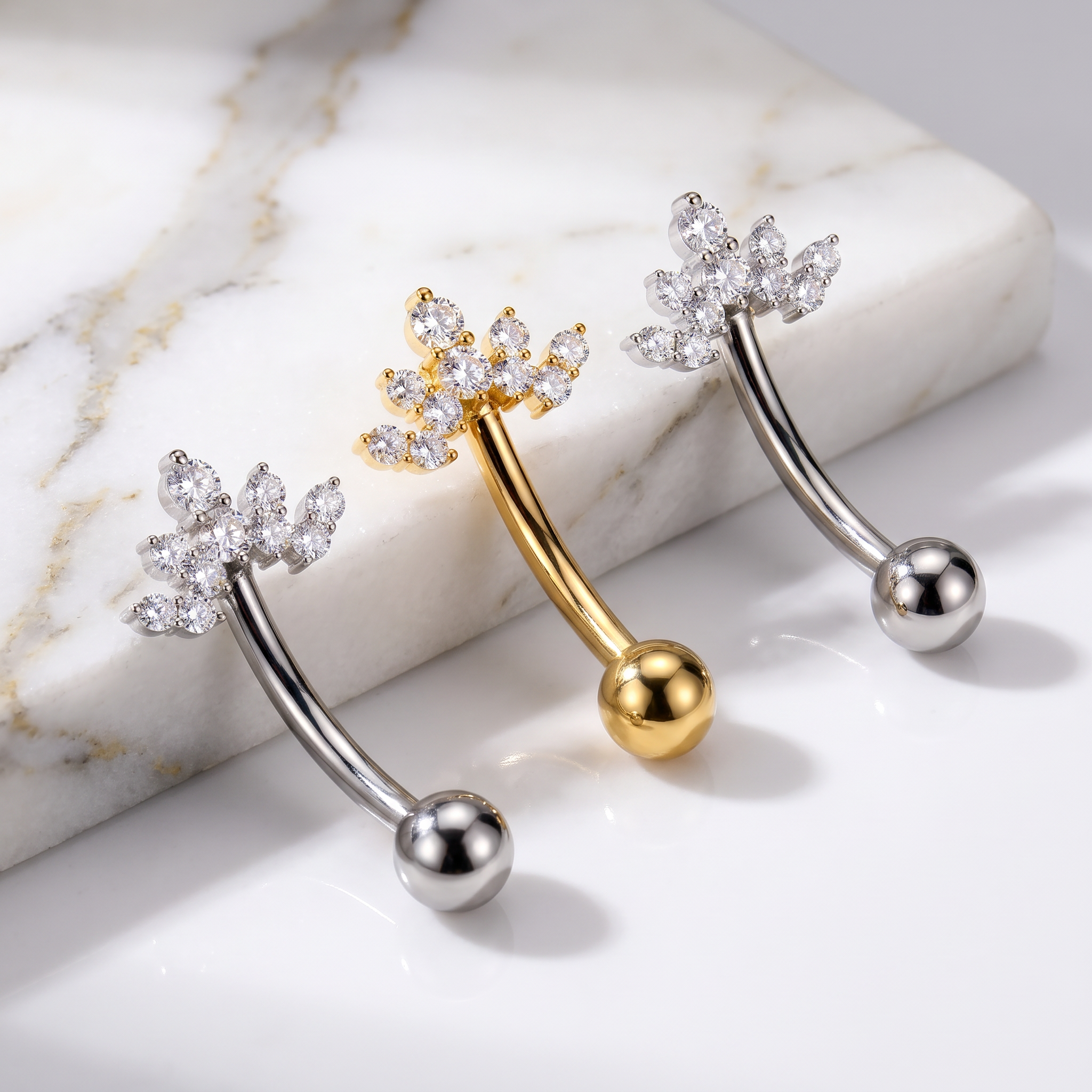 Three curved barbell belly button piercings with floral crown designs are displayed on a white marble slab. Two are silver with clear rhinestones, and one is gold with clear rhinestones.