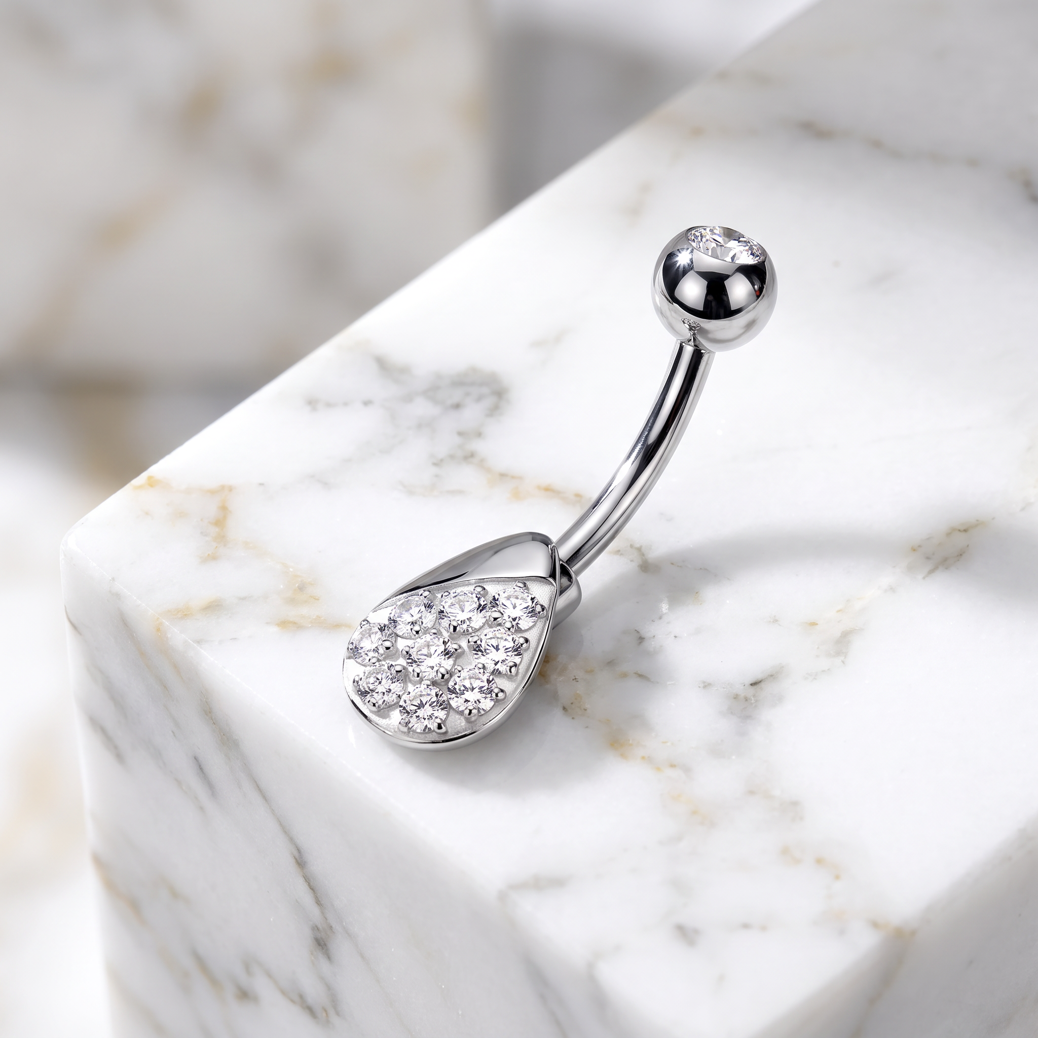 A silver-toned curved barbell belly button ring with a teardrop-shaped pendant covered in small clear stones, displayed on a white marble block.