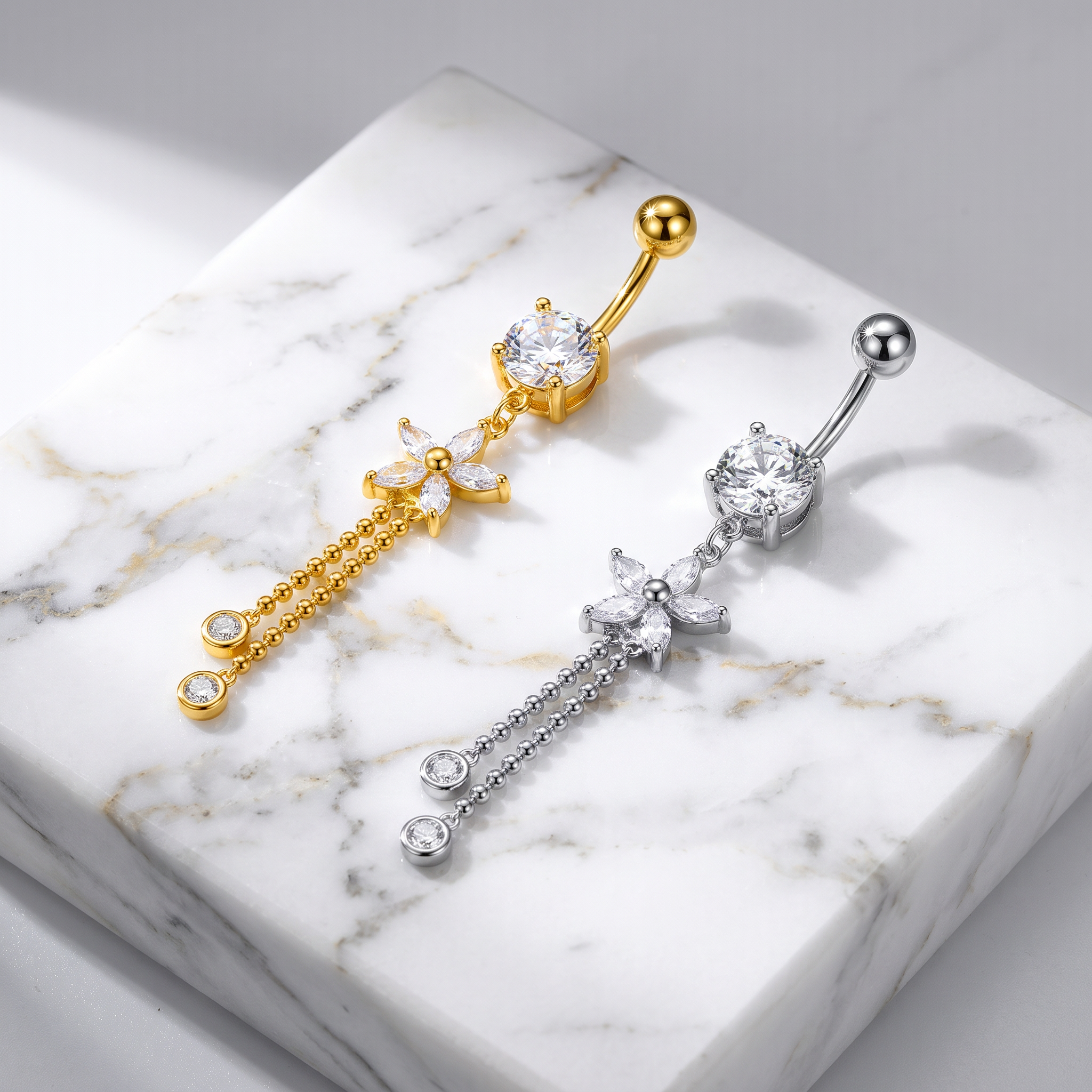 Two belly button rings with floral and dangling chain designs, one in gold and one in silver, displayed on a white marble block.