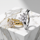Two flower-shaped rings displayed on a white marble pedestal. One ring is yellow gold with clear marquise-cut stones, and the other is silver-toned with clear marquise-cut stones.