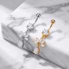 Two flower-shaped belly button piercings with teardrop drops, one in silver and one in gold, displayed on a white marble block.