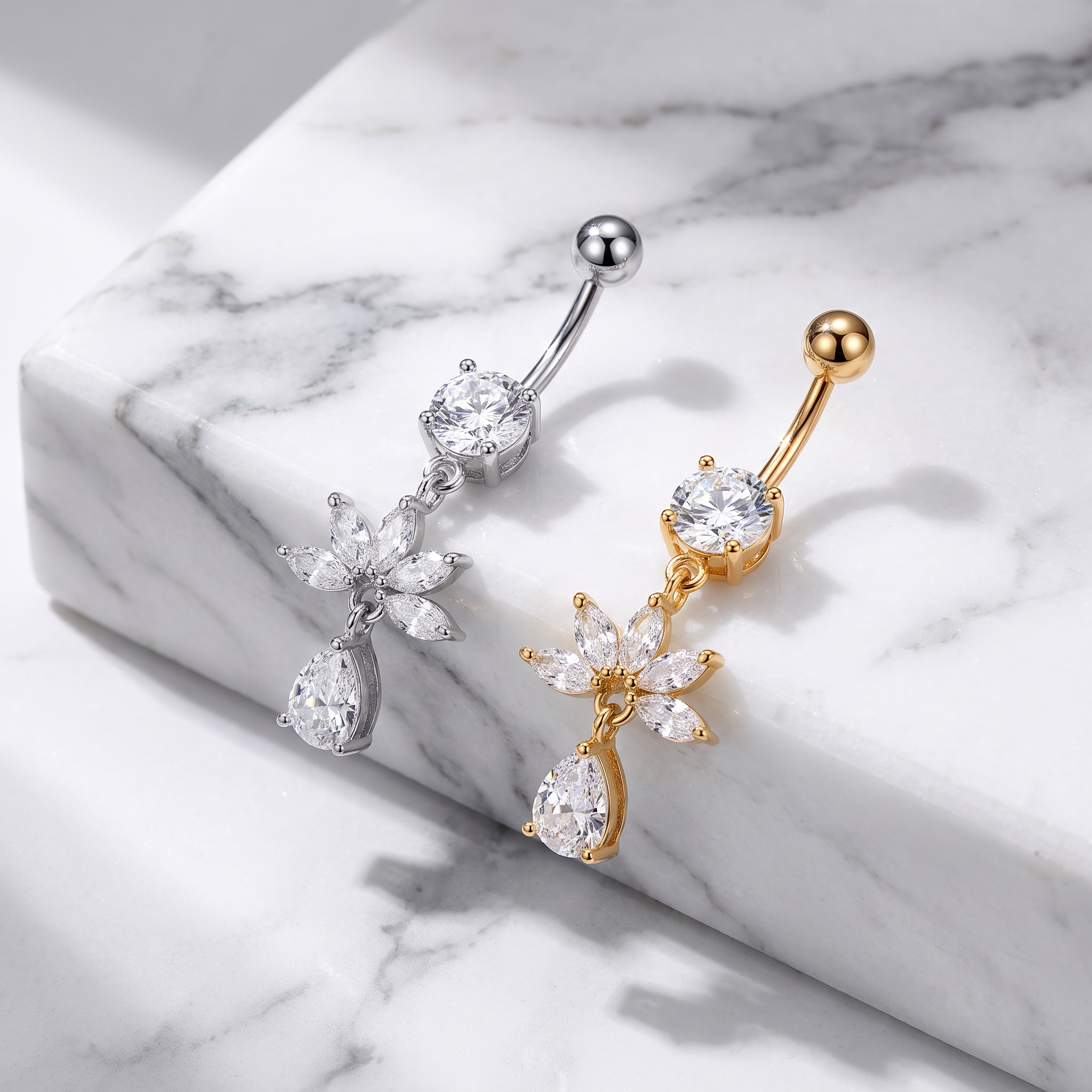 Two flower-shaped belly button piercings with teardrop drops, one in silver and one in gold, displayed on a white marble block.